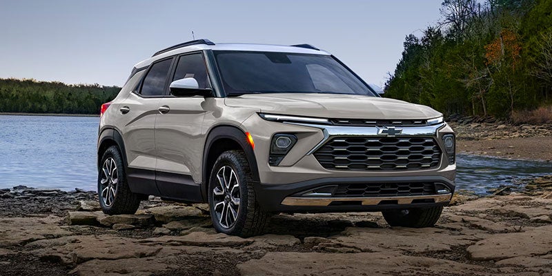 Light-colored compact SUV parked on a rocky lakeside area, shown from the front with water and trees in the background