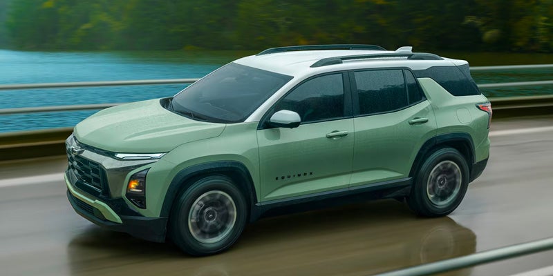 Light green compact SUV driving on a wet roadway beside a body of water, with trees visible in the background