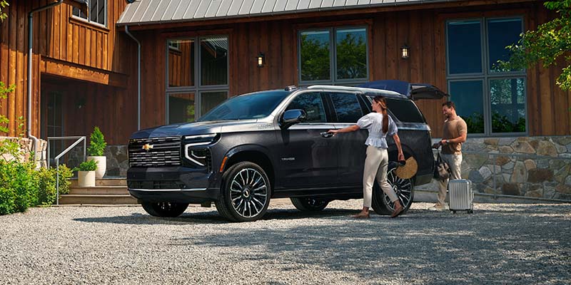 Black SUV parked in front of a wooden house while travelers load luggage into the vehicle in a residential driveway