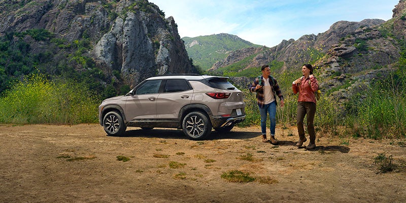 Light-colored SUV parked at a scenic mountain overlook while two people walk nearby on a dirt path surrounded by rocky cliffs and greenery