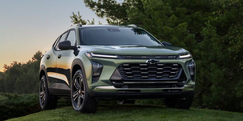 Light green compact SUV parked on a grassy area, viewed from the front three-quarter angle, with trees and a sunset sky in the background