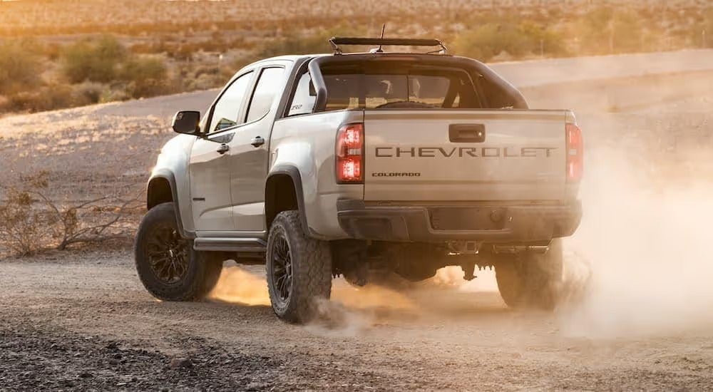 A tan 2021 Chevy Colorado ZR2, which has a factory suspension lift kit installed, is driving on a dirt road with a dust cloud rising behind it.