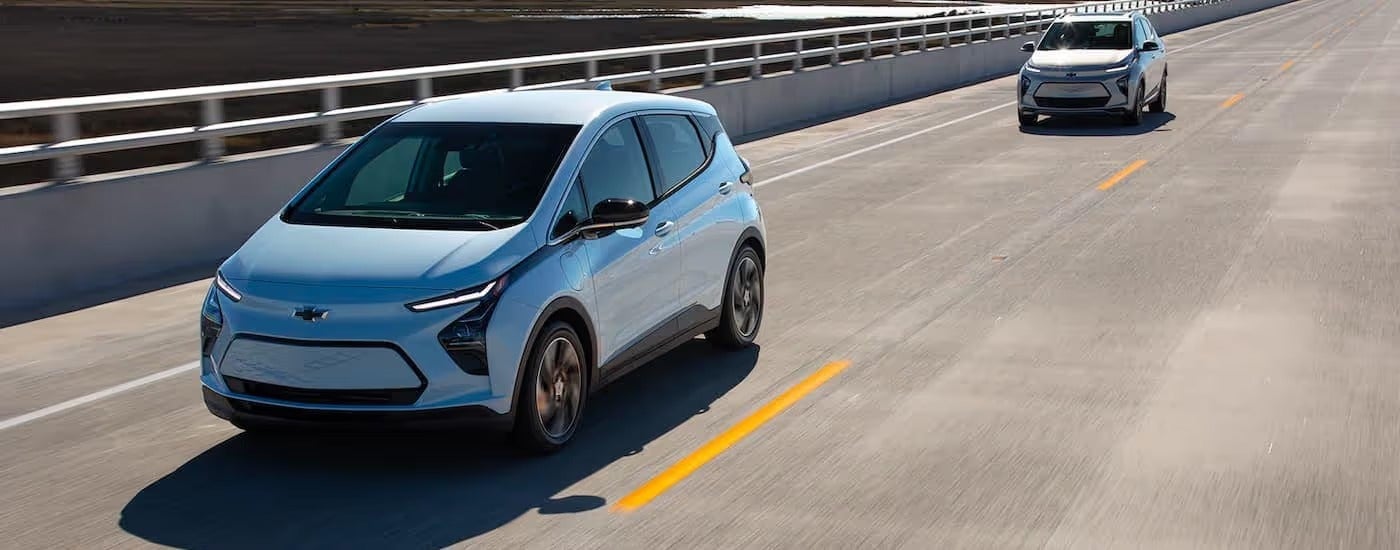 A pale blue 2022 Chevy Bolt EV and a silver 2022 Bolt EUV are driving down an empty highway.
