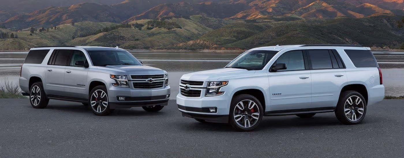 A silver Chevy Suburban and white Chevy Tahoe in front of a lake and mountains