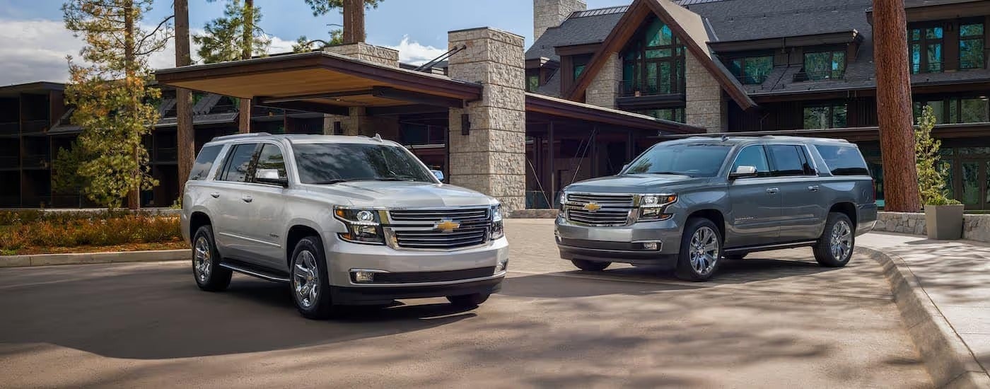 A silver 2020 Chevy Tahoe is parked next to a grey Suburban Premier Plus in a cabin driveway.