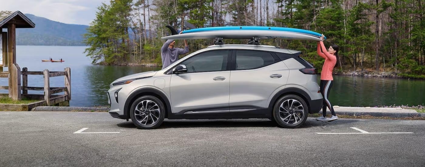 A silver 2022 Chevy Bolt EUV is shwon from the side in front of a lake while a couple gets paddle boards off the roof.