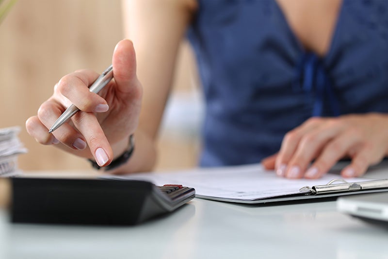 Person using a calculator while reviewing paperwork at a desk, holding a pen and entering figures