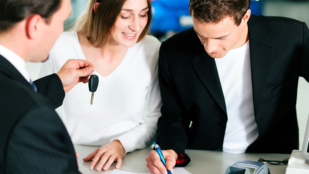Car buyer signing documents while another person hands over a vehicle key during a dealership transaction