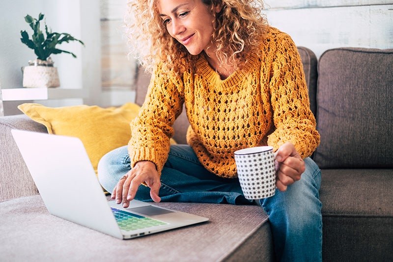 Person sitting on a sofa using a laptop while holding a mug, with a living room setting and decorative pillows in the background