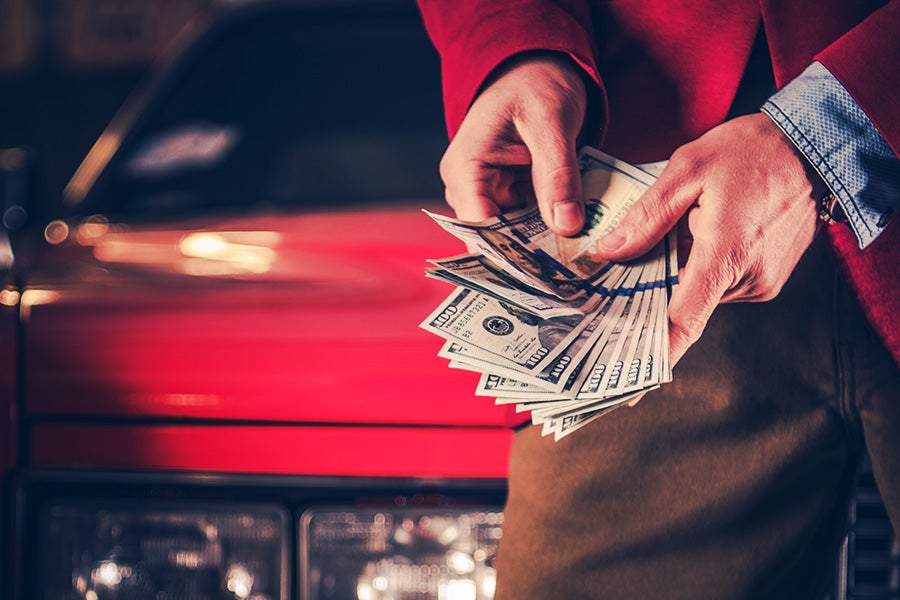 Person holding and counting U.S. dollar bills in front of a red vehicle, with the car's front end blurred in the background